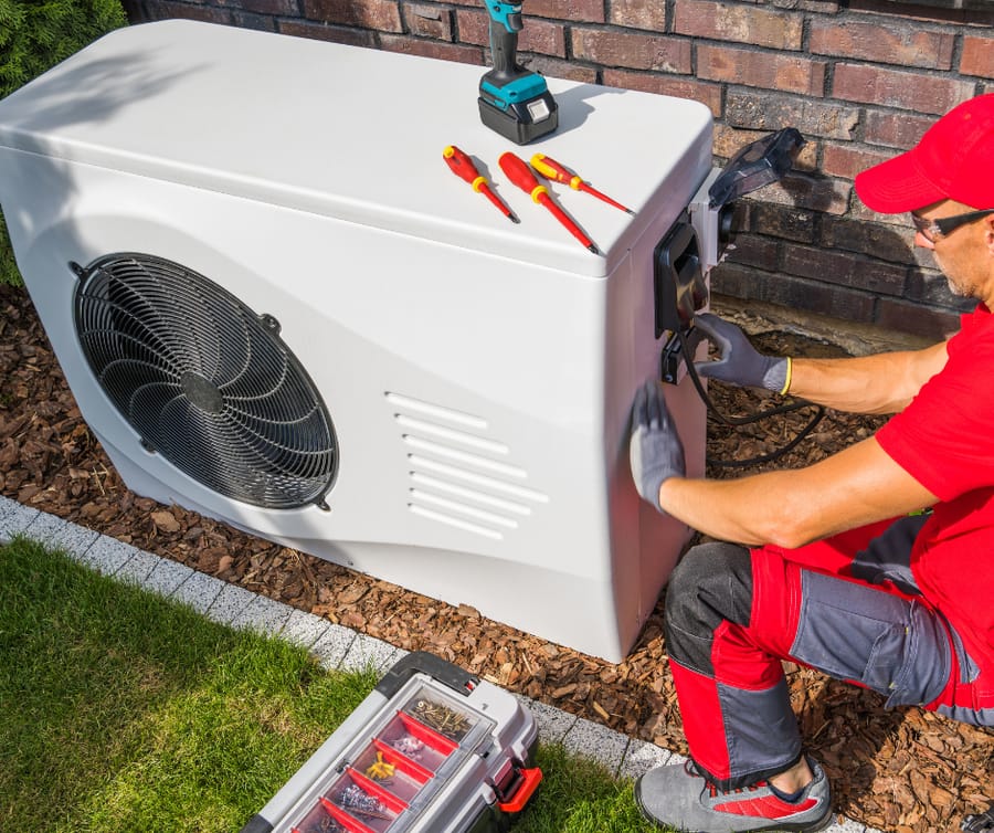 HVAC technician performing service on heat pump at a residential home; his tools are on top of the heat pump and on the ground as he works