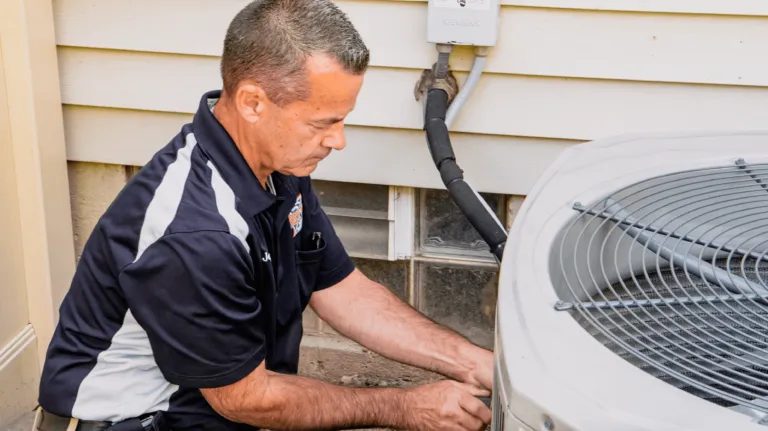 Technician in a dark polo shirt crouches beside an outdoor air conditioner unit, working on it.