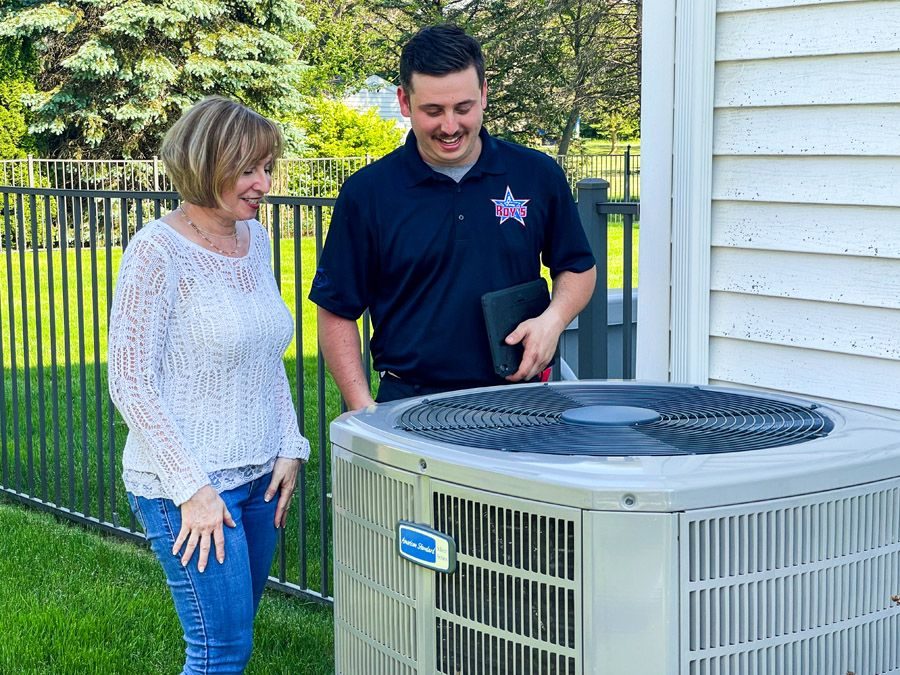 A technician explaining an outdoor air conditioner unit beside a smiling homeowner in a backyard fence setting.