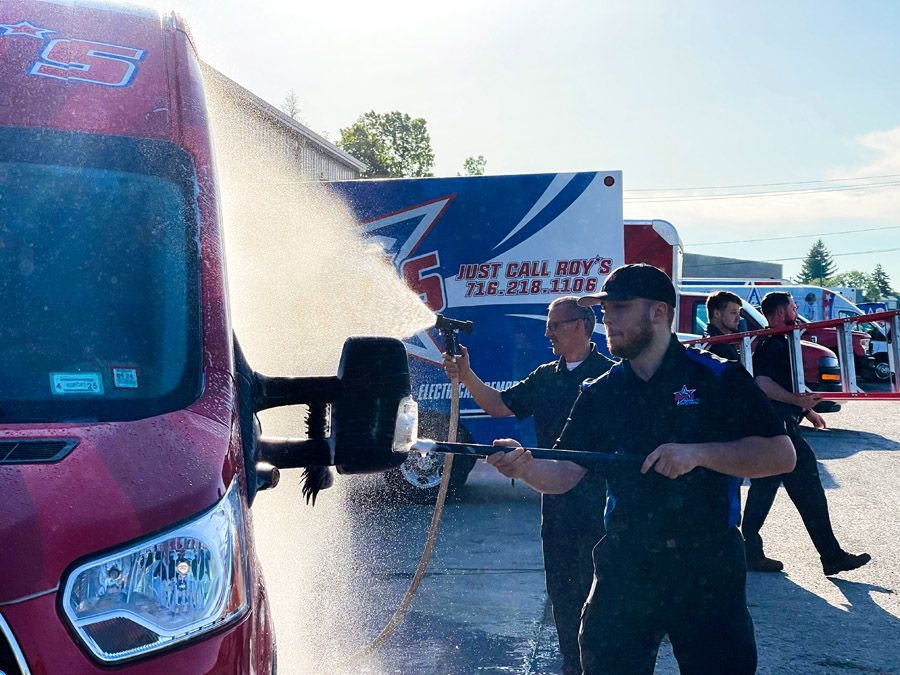 Two workers in dark uniforms use a high-pressure hose to wash a red delivery truck at an event.