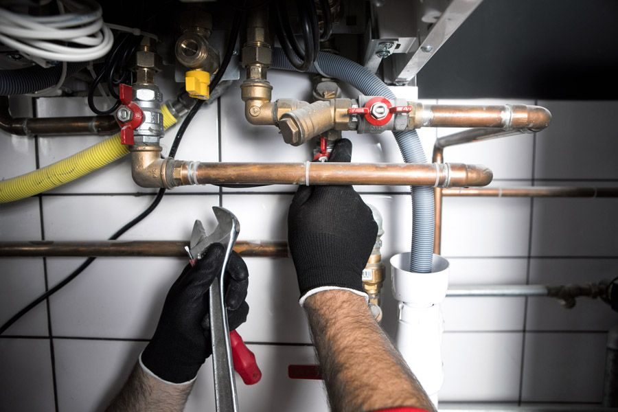 Plumber wearing black gloves tightening copper pipes and valves under a sink with a wrench.