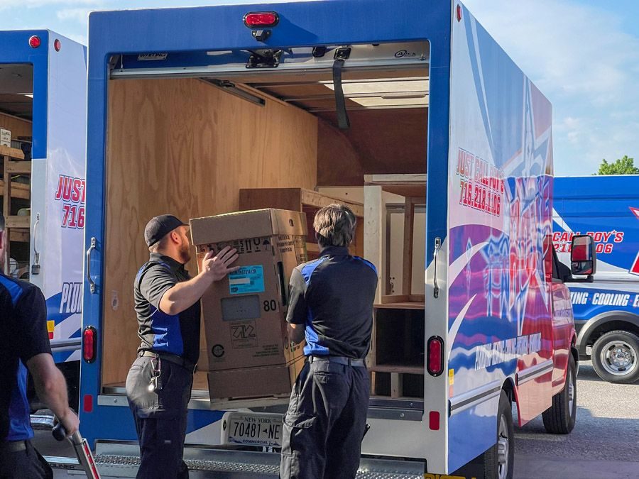 Two workers in uniforms load a large cardboard box into the back of a blue moving van with plywood interior while another truck is visible in the background.