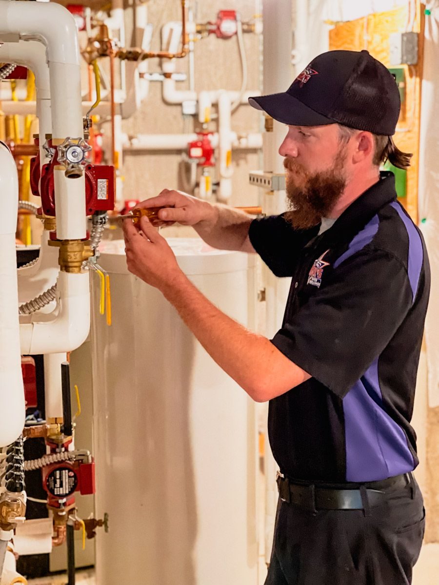 Maintenance technician in a black and purple uniform adjusts a red valve on industrial piping in a utility room.