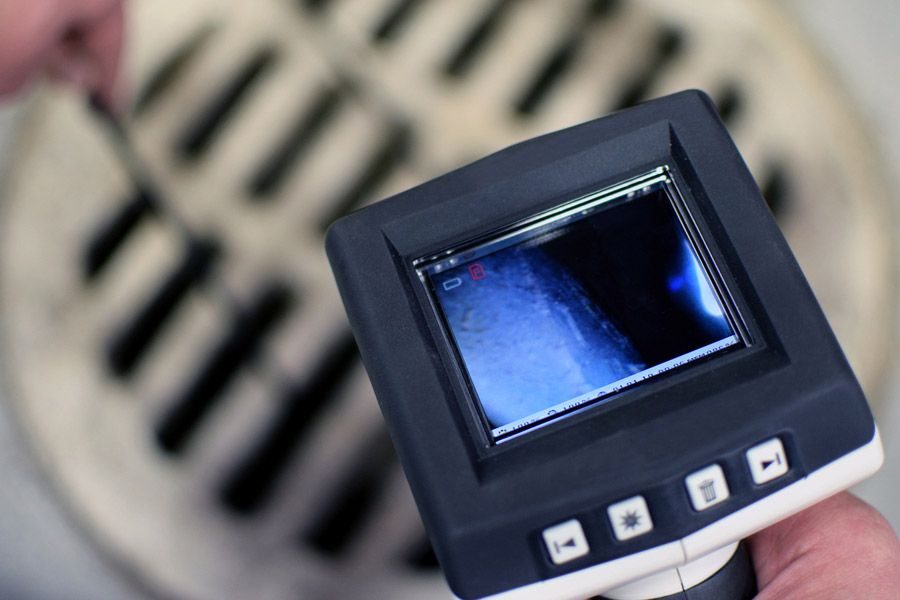 Handheld inspection camera with a small screen showing a blue image over a metal drain grate in the background
