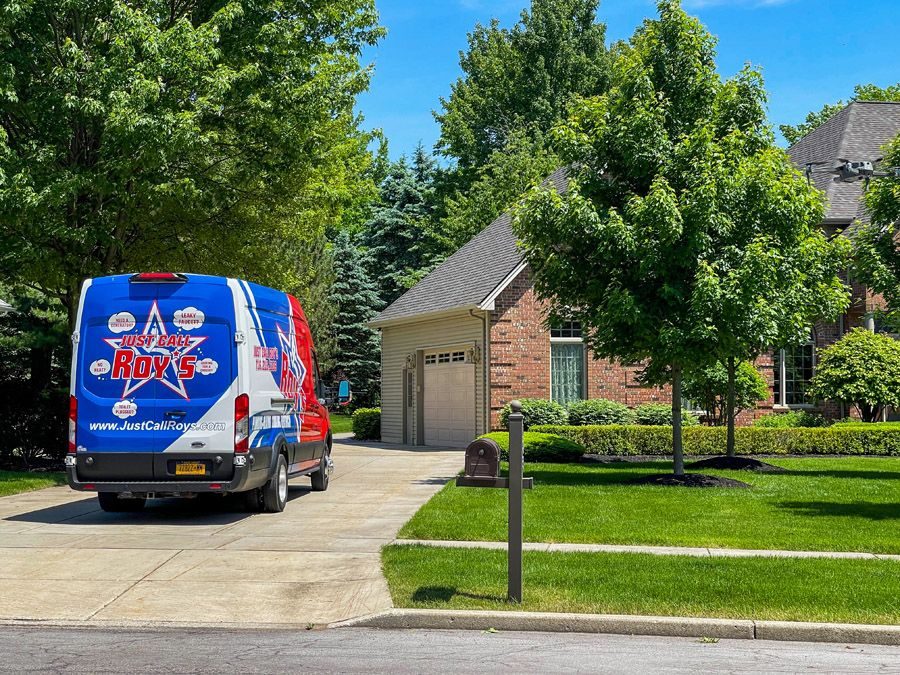 Branded delivery van parked in a suburban driveway beside a brick house with a two-car garage, surrounded by green trees and a manicured lawn.