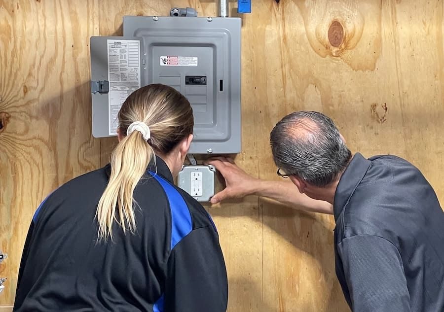 Two technicians inspect a wall-mounted electrical panel and outlet in a wood-paneled workshop, with one person pointing at the outlet cover.