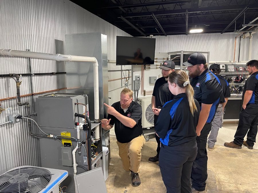 Instructor kneeling and explaining HVAC equipment to a group of technicians during a training session in a workshop.