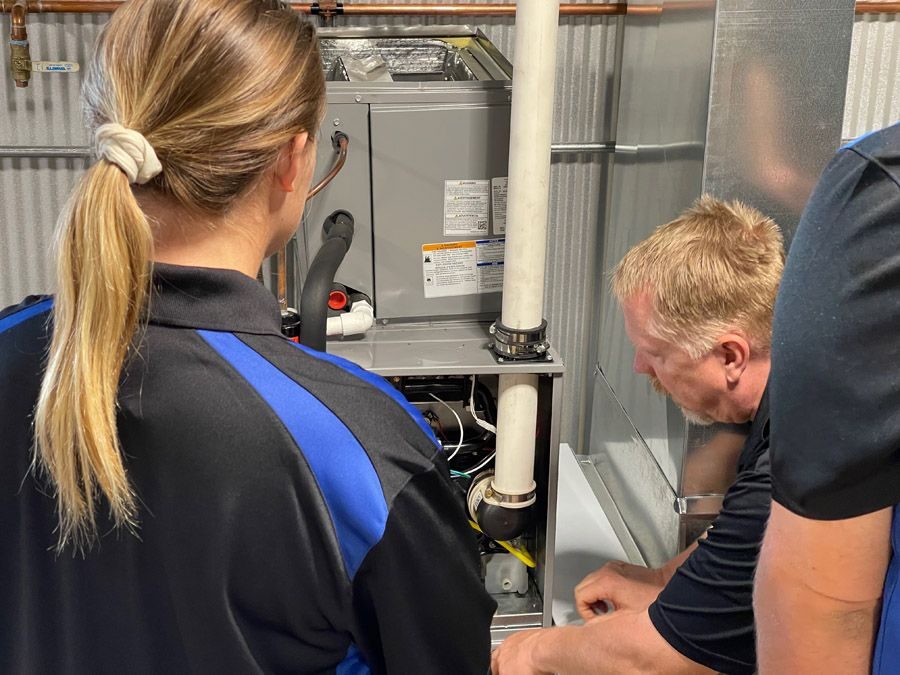 Two technicians inspect and repair an open furnace cabinet in a utility room; tools and white PVC pipe visible nearby
