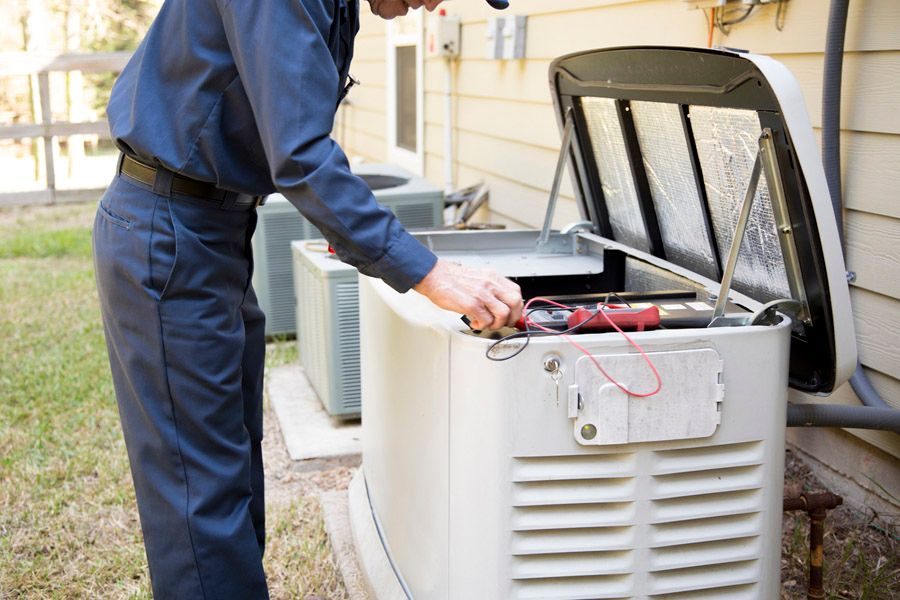 HVAC technician in a blue uniform testing electrical wires inside an outdoor air conditioner condenser unit with a multimeter.