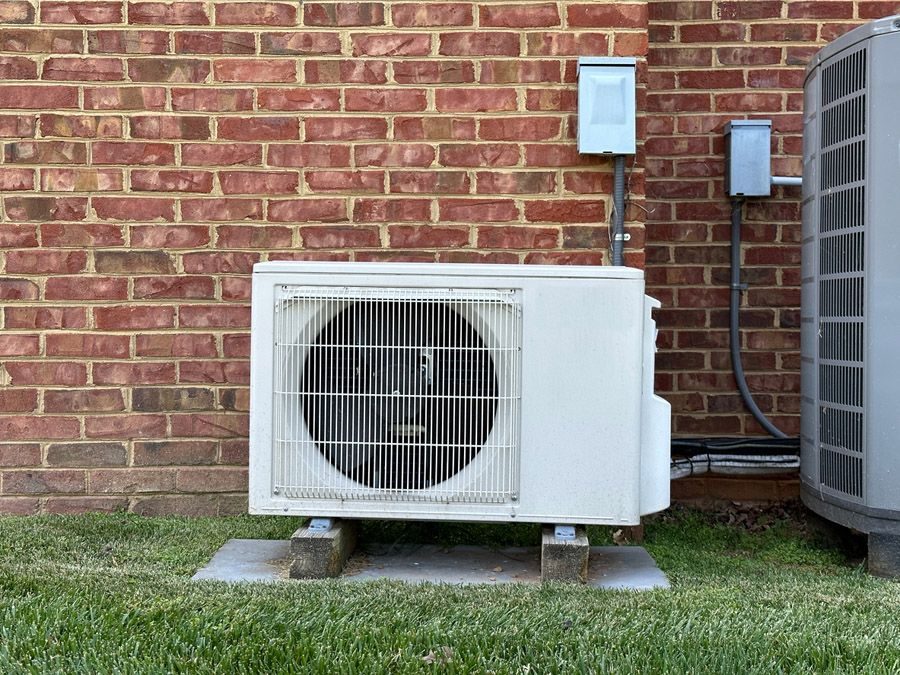 Outdoor air conditioner condenser with protective grille mounted on concrete blocks against a brick wall, with a nearby unit and wiring.