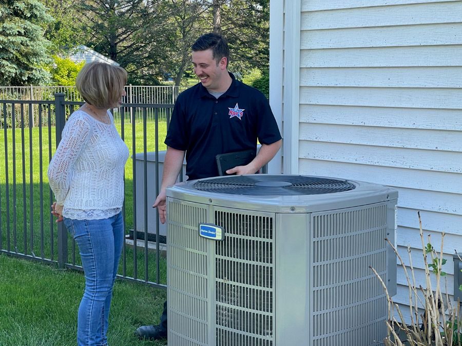 Two adults standing beside a large outdoor air conditioning condenser, smiling and talking in a sunny backyard next to a white house wall.