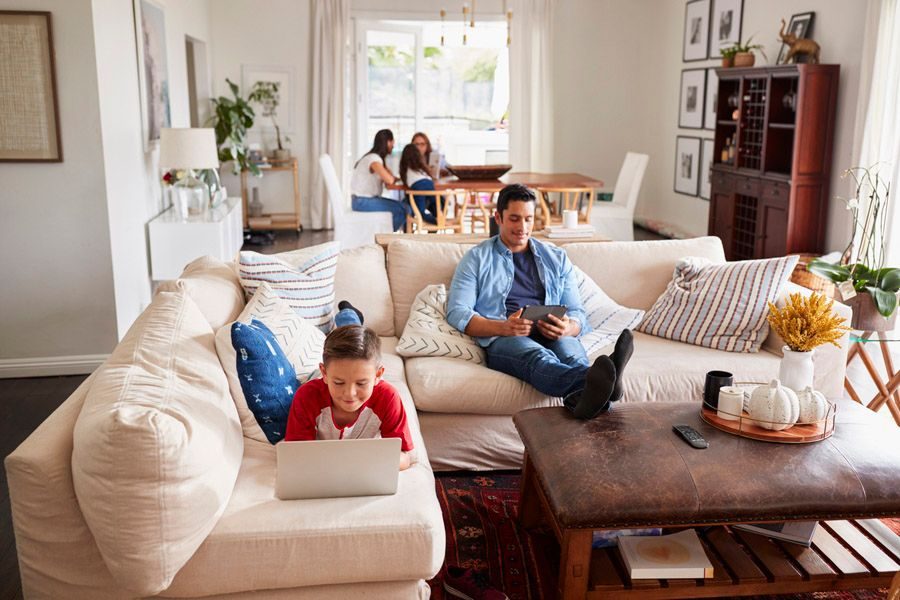 Family relaxing in a bright living room: a man on a couch using a tablet while a child on the sofa uses a laptop, with a dining area in the background.