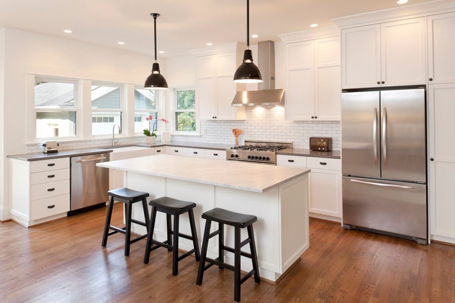 Bright white kitchen with an island, three black stools, and stainless steel fridge and oven under pendant lights.