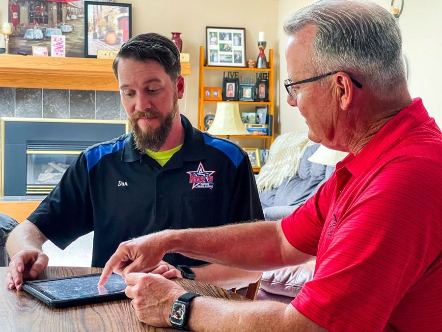 Two men sit at a wooden table looking at a tablet; one wears a black Roy's shirt with name Dan, the other in a red polo.