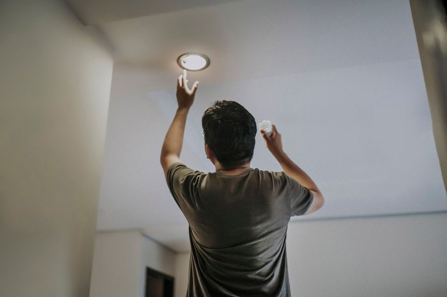 Man seen from behind reaching up to a ceiling light, holding a cloth in the other hand in a indoor room.