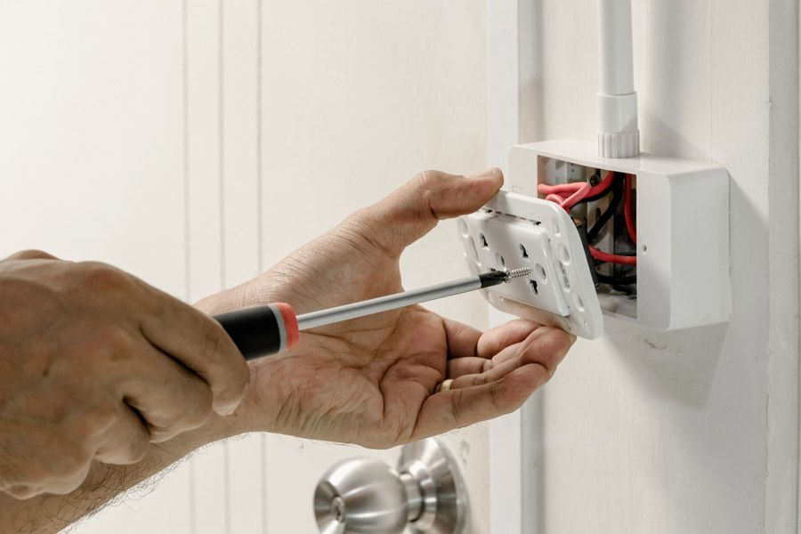 Person using a screwdriver to secure a wall-mounted electrical outlet cover, with exposed wires visible behind the plate