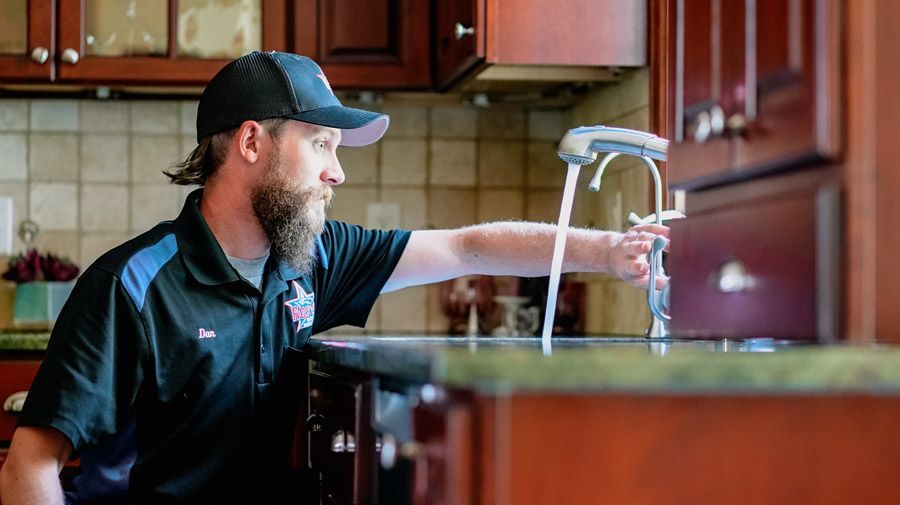 Bearded man in a cap and polo shirt reaches toward a kitchen sink faucet, inspecting the chrome fixture in a home kitchen.