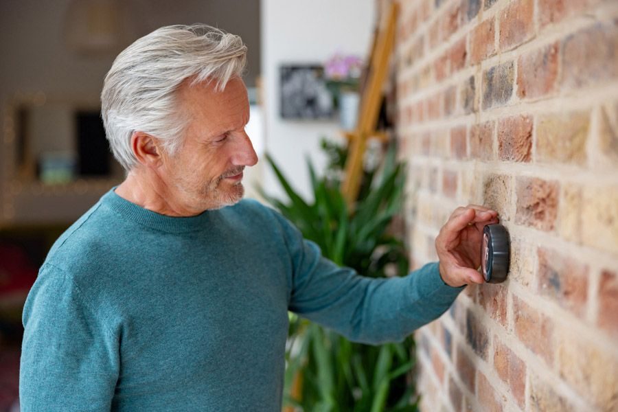 Older man in a teal sweater presses a round doorbell on a brick wall inside a home.