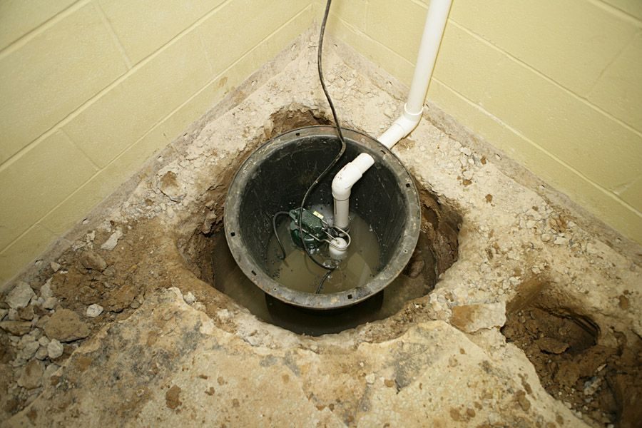 Sump pump basin in a dug hole in a basement floor with PVC pipes and electrical cords partially submerged in dirty water.