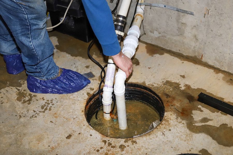 Plumber in blue sleeves and shoe covers works on an open basement floor drain with white PVC pipes submerged in water.