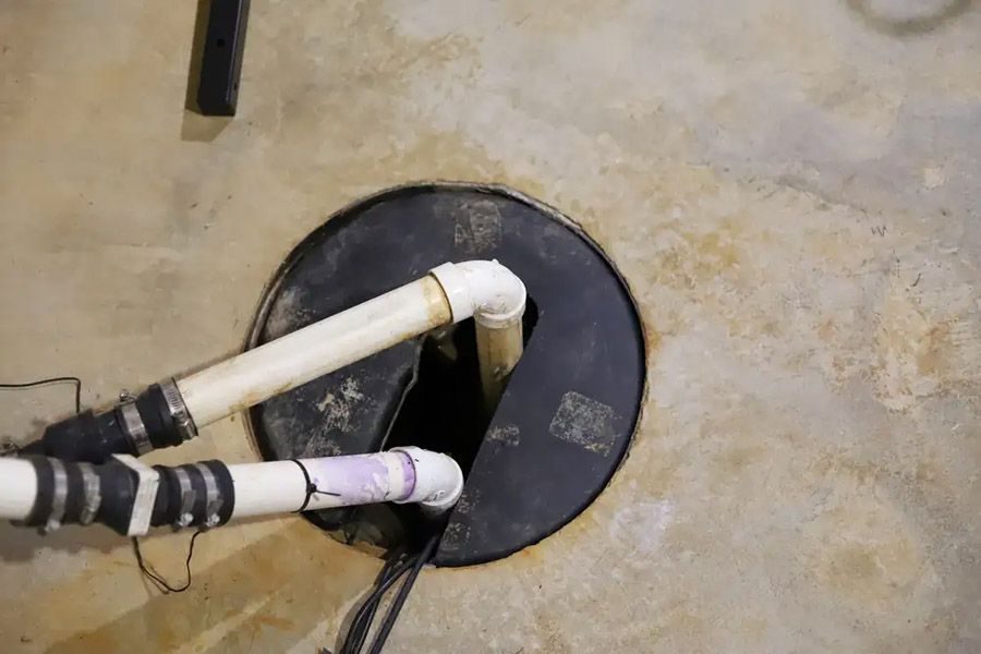 Floor drain/cleanout with white PVC pipes and elbow joints emerging from a circular black access cover in a concrete floor.