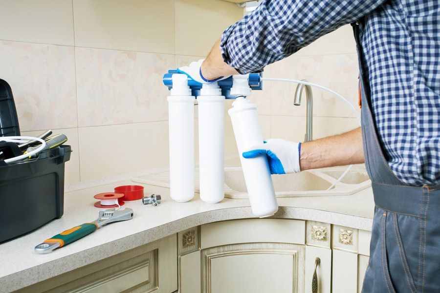 A person wearing gloves replaces water filter cartridges on an under-sink water filtration system in a kitchen.