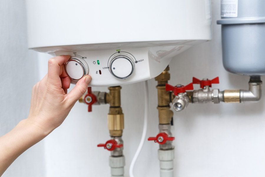 Person turning a dial on a wall-mounted water heater, with red valve handles and pipes in the background.