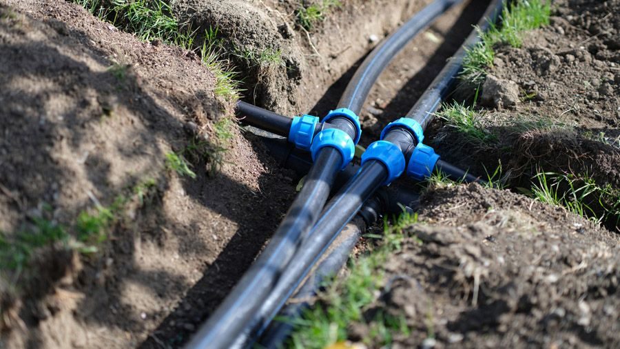Black irrigation hoses with blue connectors laid in a trench in dirt and grass near the surface of the soil.