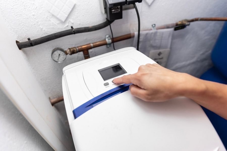 Hand pressing the start button on a top-loading washing machine in a utility room with exposed pipes in the background.