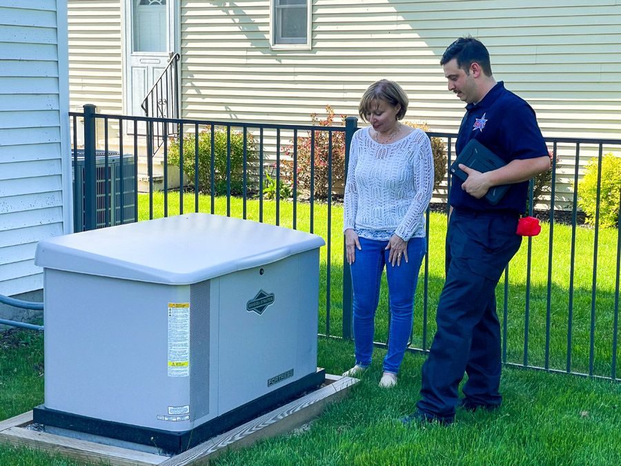 Service technician in dark uniform and a homeowner inspect a large outdoor standby generator on a wooden pad in a fenced backyard.