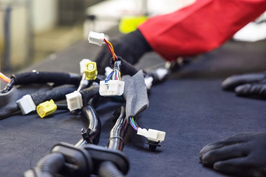 Close-up of a car wiring harness with white connectors and multicolored wires on a workbench; gloved hands nearby indicating repair work