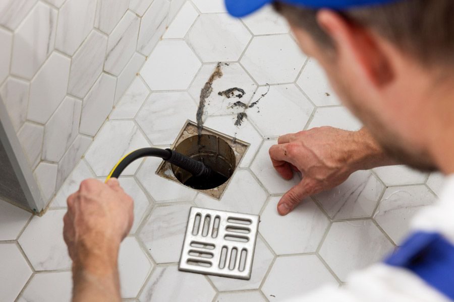 Plumber inserts a drain snake into a square floor drain in a tiled bathroom.