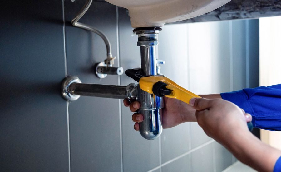 Person using a pipe wrench to tighten a shiny chrome sink drain under a dark wall panel.