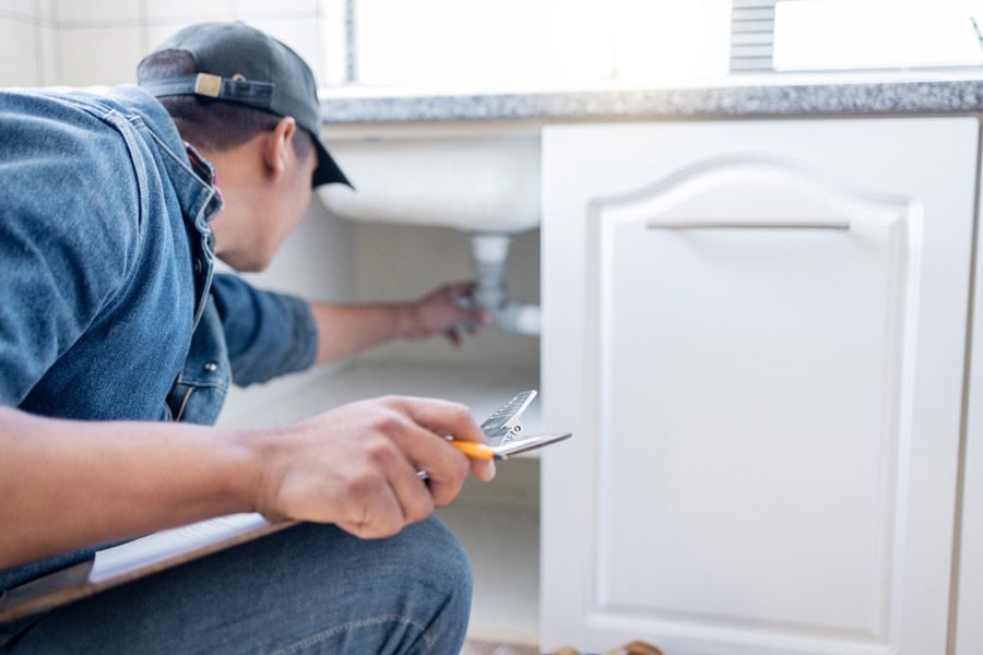 A repair technician crouches under a kitchen sink, inspecting pipes and holding a ruler and utility knife