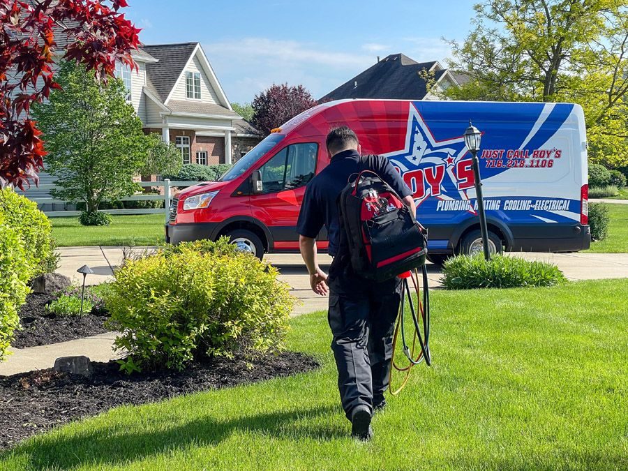 A technician with a backpack carrying hoses walks across a green lawn toward a red‑and‑blue service van parked in a suburban driveway.